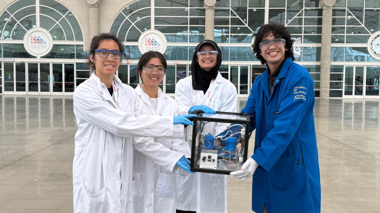 Four students in lab coats hold a cube structure in front of a building with windows